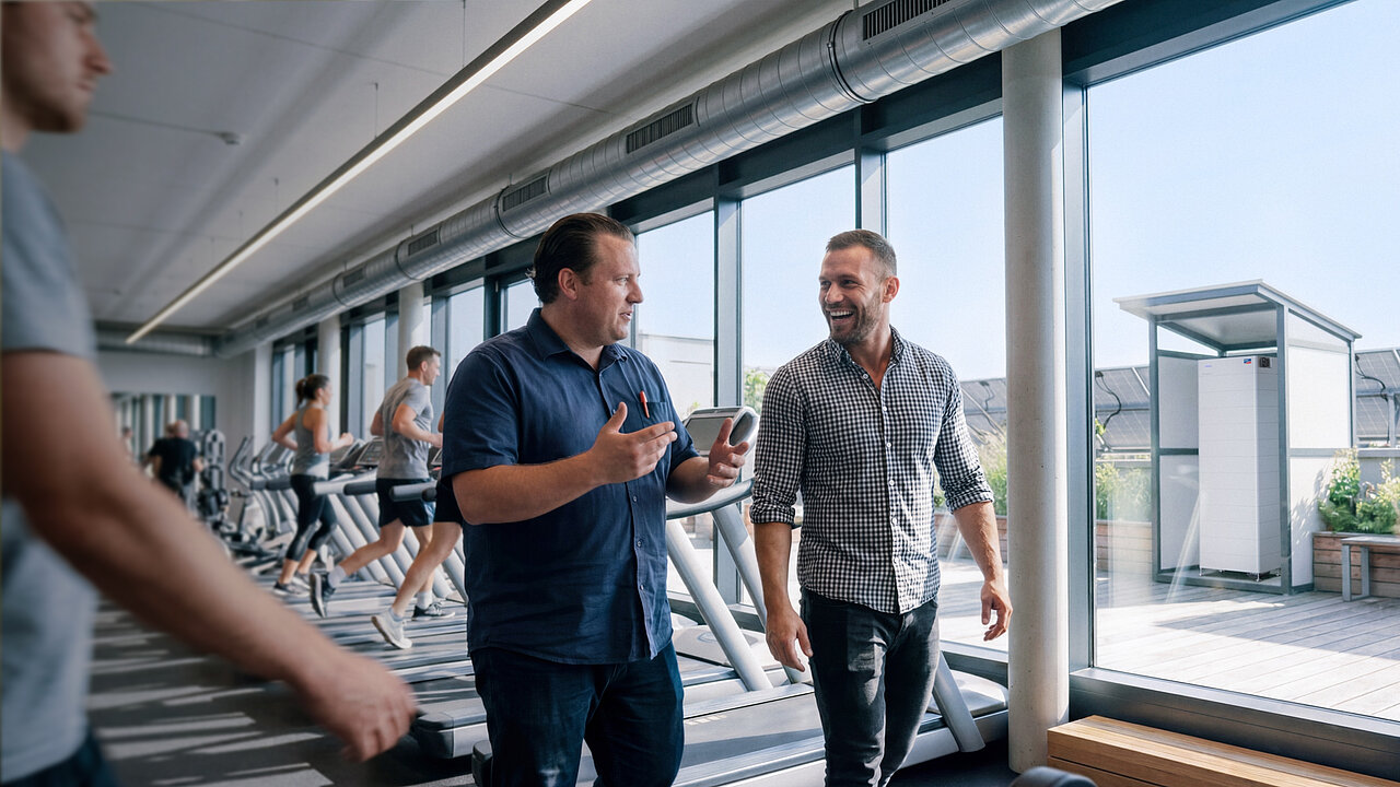 Two men—deep in conversation—walk past a row of treadmills set up in front of a large window. Outside the window stands an SMA Storage M.