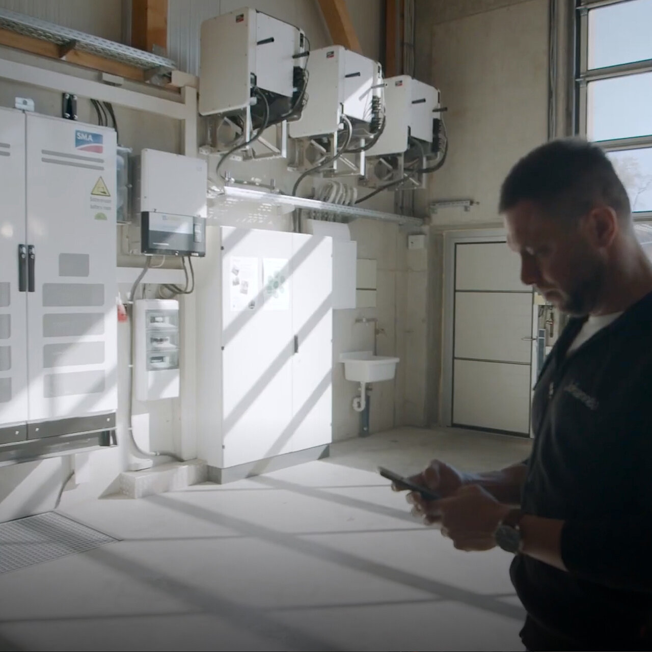 A large hall with several SMA Sunny Tripower CORE1 units mounted on the wall, an SMA control cabinet, and another fuse box; in front of them stands a man looking at his cell phone.
